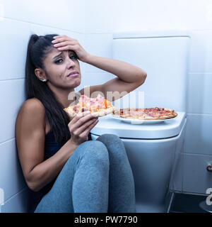 Attraente giovane e triste bulimic giovane donna di sentirsi colpevole e malati di mangiare mentre è seduto sul pavimento accanto al bagno in disturbi alimentari anoressia Foto Stock
