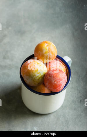 Pila di colorati multicolore di appena raccolto le prugne Mirabelles rosso verde giallo in smalto bianco mug su nero grigio pietra dello sfondo. Autunno raccolto autunnale Foto Stock