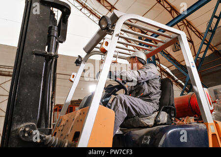 Uomo alla guida di un carrello elevatore a forche attraverso un deposito in una fabbrica. driver in uniforme e casco protettivo. Il concetto di logistica e di storage Foto Stock