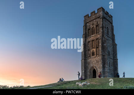 Una piccola folla si riuniscono per guardare il sole che tramonta accanto alla Parrocchia di San Michele torre sulla sommità di Glastonbury Tor in Somerset. Foto Stock