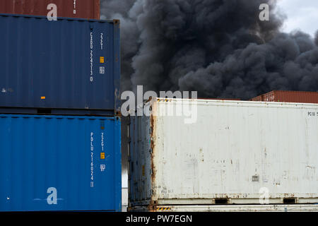 Vista astratta di contenitori di spedizione con pennacchi di fumo tossico da un incendio industriale sollevarsi verso il cielo. Foto Stock