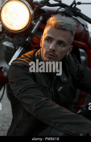 Elegante ritratto di una brutale hipster modello uomo in un elegante giacca militare si trova nei pressi di una motocicletta con luce di sera Foto Stock