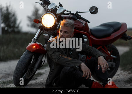 Elegante bel giovane uomo brutale in una zona alla moda di camicia cachi si trova nei pressi di un motociclo nel campo Foto Stock