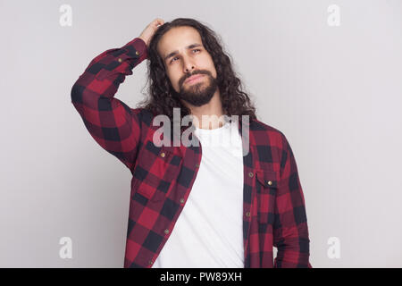 Confuso uomo bello con barba nera e lunghi capelli ricci in casuale a scacchi in maglietta rossa in piedi, di pensare e di guardare lontano con faccia pensieroso . in Foto Stock
