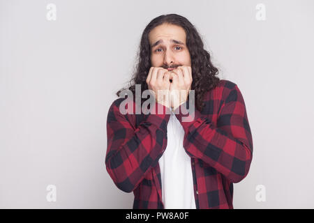 Ritratto del sistema nervoso o turbare l uomo con la barba nera e lunghi capelli ricci in stile casual, a scacchi maglietta rossa in piedi guardando la fotocamera e bitting chiodo Foto Stock