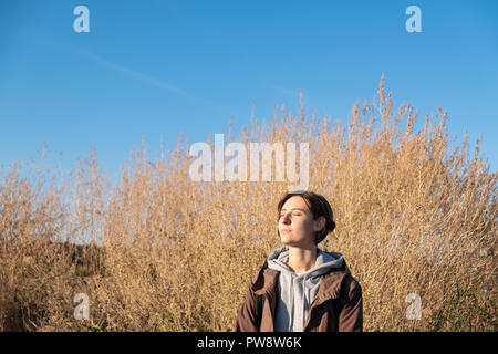 Giovane donna gode del sole autunnale. Femmina in parka all'aperto in un prato sul luminoso pomeriggio soleggiato Foto Stock