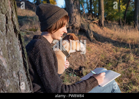 Escursionista la scrittura di un diario di viaggio nella splendida natura. La donna rivolta verso il sole di sera prende appunti in un blocco note, cane si siede accanto a lei Foto Stock