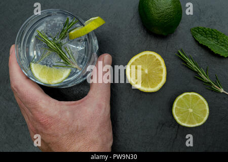 Mano che regge un bicchiere di gin tonic con ghiaccio e calce su sfondo scuro Foto Stock