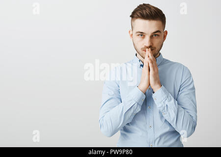 Smart creative ambizioso uomo bello con setola in elegante camicia formale tenendo le mani nel pregare sulla bocca nella Genius pongono pensando o rendendo importante decisione preoccupante oltre il muro grigio Foto Stock