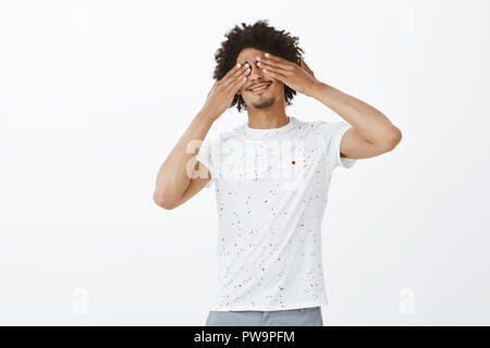 Carino felice e joyfuly fidanzato chiudendo gli occhi e in attesa di sorpresa b-giorni di festa. Carefree bello modello maschile con la pelle scura e i baffi, che copre la vista con palme e sorridente con gioia Foto Stock