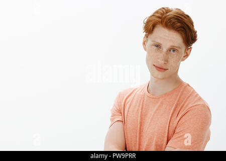 Guy può utilizzare il suo fascino per sedurre qualsiasi donna. Ritratto di bello fiducioso e spensierato redhead studente maschio con lentiggini in t-shirt, tenendo le mani incrociate e guardando con leggero sorriso Foto Stock