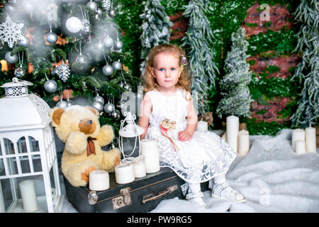 Incantevole piccola ragazza in un abito bianco seduto vicino a un albero di Natale su una valigia accanto alle candele e un orso di peluche Foto Stock