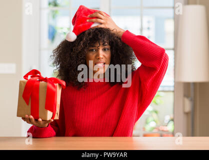 African American donna tenendo presente che indossa il natale Red Hat ha sottolineato con la mano sulla testa, sconvolto con vergogna e la sorpresa di fronte, arrabbiato e vanificare Foto Stock