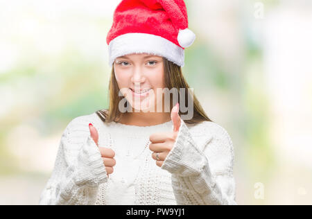 Giovani caucasici bella donna con cappello a Natale su sfondo isolata facendo felice Thumbs up gesto con la mano. Recante approvazione di espressione in cerca di un Foto Stock