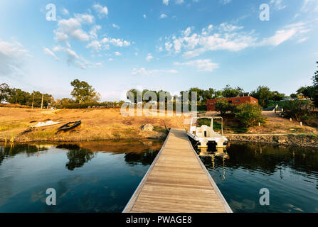 Pontile in legno con barca ormeggiata e riflessioni sul lago di acqua con un cielo blu con nuvole in background Foto Stock