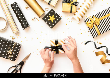 Vista dall'alto di mani femminili di incarto scatole regalo, sparsi in materiali di imballaggio in vari nero, bianco e disegni dorati. Un concetto di Natale, nuovo sì Foto Stock