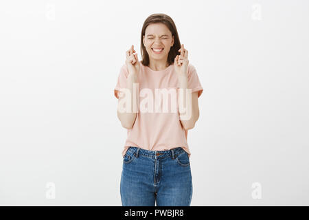 Excited donna allegra in abito casual, chiudendo gli occhi e sorridendo ampiamente mentre si incrociano le dita e rendendo desidera, sognare o sperando in qualcosa di vero, di pregare per il buon risultato in esame Foto Stock