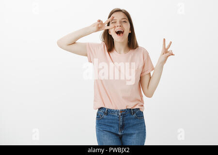 Ritratto di spensierata ragazza europea in vestiti alla moda, che mostra la vittoria o gesto di pace over eye, winking e sorridente largamente, sensazione di grande divertendosi con gli amici, appendere fuori felicemente Foto Stock