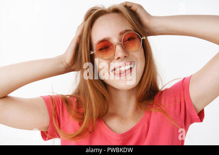 Ragazza elegante nel quartiere alla moda di occhiali da sole e t-shirt rosa holding hands diffusione sulla testa winking, spuntavano lingua felicemente e sorridente dalla gioia e rilassata emozioni divertendosi, rilassante oltre il muro grigio Foto Stock