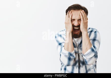 L'uomo sensazione emozioni dolorose, urlando, testimoniando la morte del caro amico, essendo miserabili e devastato, tenendo le palme sulla testa, urla di dolore e di stress, infelice permanente oltre il muro grigio Foto Stock