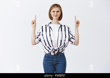 Suoni di sospetto. Ritratto di certi moderni smart femmina con capelli biondi in camicia a righe, smirking dal dubbio e accigliata mentre rivolto verso l'alto con le dita indice, sensazione di dubbia Foto Stock