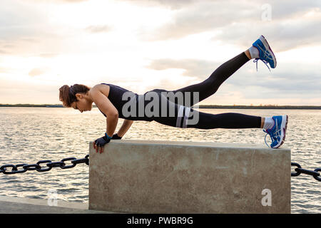 Una donna attraente in abbigliamento sportivo facendo esercizi sportivi in natura contro il tramonto e il fiume di Amur, ama la ginnastica, massaggia le gambe. Attivo giovane ragazza impegnate nello sport, conduce uno stile di vita sano. Foto Stock