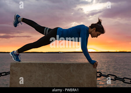 Una donna attraente in abbigliamento sportivo facendo esercizi sportivi in natura contro il tramonto e il fiume di Amur, ama la ginnastica, massaggia le gambe. Attivo giovane ragazza impegnate nello sport, conduce uno stile di vita sano. Foto Stock