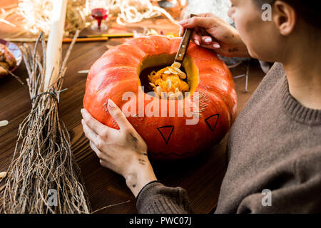 Close-up di concentrato di donna in maglia seduta a tavola con la scopa e scooping fuori i semi di zucca mentre rendendo jack-o-lantern in officina Foto Stock
