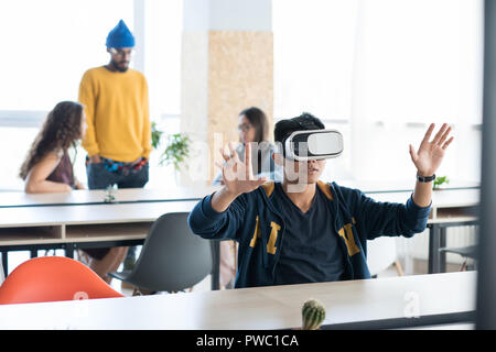 Contenuto bel giovane maschio asiatici gli sviluppatori di app in abbigliamento casual seduta a tavola e gesticolando mani mentre utilizzando la realtà virtuale simulatore di cowor Foto Stock