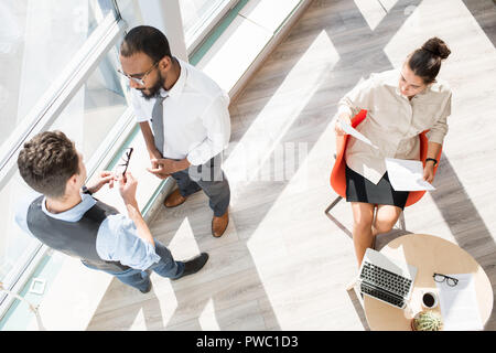 Direttamente al di sopra di vista del moderno business persone in convenzionale lavora in ufficio: grave multi-etnico imprenditori a discutere di progetto mentre concentrata Foto Stock