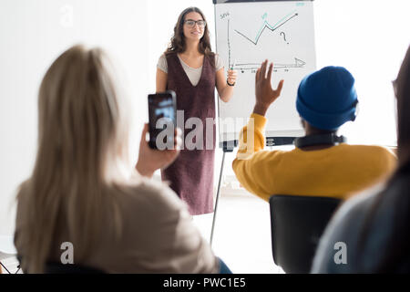 Allegro fiducioso femmina sales manager in bicchieri in piedi alla lavagna con disegno grafico e tenendo un seminario per gli studenti, ella rispondendo alle domande Foto Stock