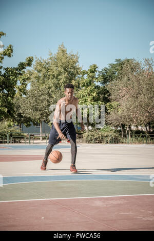 Uno afroamerican giovane senza tshirt è giocare a basket in un parco a Madrid durante il periodo estivo a mezzogiorno. Egli sta facendo un salto impressionante per effettuare una connessione DUN Foto Stock