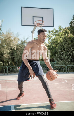 Uno afroamerican giovane senza tshirt è giocare a basket in un parco a Madrid durante il periodo estivo a mezzogiorno. È lui che consiste nel far rimbalzare la palla sotto il cestello. Foto Stock
