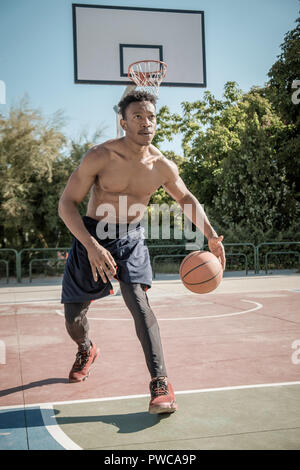 Uno afroamerican giovane senza tshirt è giocare a basket in un parco a Madrid durante il periodo estivo a mezzogiorno. È lui che consiste nel far rimbalzare la palla sotto il cestello. Foto Stock