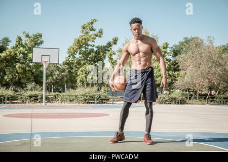 Uno afroamerican giovane senza tshirt è giocare a basket in un parco a Madrid durante il periodo estivo a mezzogiorno. Egli è in piedi e loooking presso la fotocamera Foto Stock