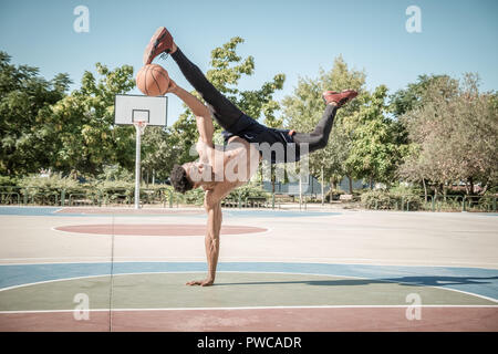 Uno afroamerican giovane senza tshirt sta facendo in equilibrio con una palla da basket in un parco a Madrid durante l'estate a mezzogiorno. Foto Stock