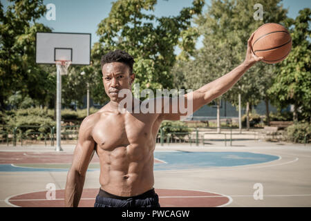 In prossimità di uno afroamerican giovane senza tshirt giocare a basket in un parco a Madrid durante il periodo estivo a mezzogiorno. Egli è in piedi e tenendo il b Foto Stock