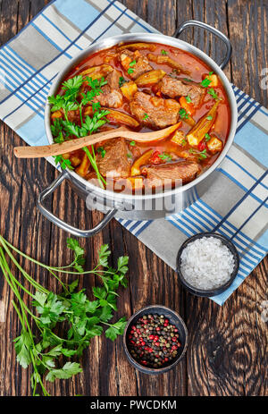 Deliziosa carne di manzo e Okra stufare in una casseruola di metallo con cucchiaio, su un vecchio tavolo di legno, vista verticale dal di sopra, close-up Foto Stock