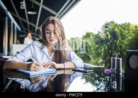 Grave studentessa in occhiali concentrata sul piano di scrittura nel blocco note mentre l'invio di messaggi sul cellulare, pensieroso hipster ragazza in eyewear rilevando Foto Stock