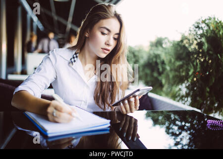 Grave studentessa in occhiali concentrata sul piano di scrittura nel blocco note mentre l'invio di messaggi sul cellulare, pensieroso hipster ragazza in eyewear rilevando Foto Stock