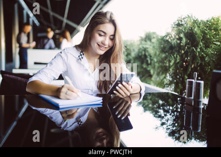 Grave studentessa in occhiali concentrata sul piano di scrittura nel blocco note mentre l'invio di messaggi sul cellulare, pensieroso hipster ragazza in eyewear rilevando Foto Stock