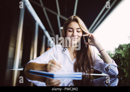 Studente elegante nel quartiere alla moda di hat parlando con un amico sul telefono e rilevando alcuni rapporti in Blocco note in seduta cafè all'aperto.Creative hipster ragazza writi Foto Stock