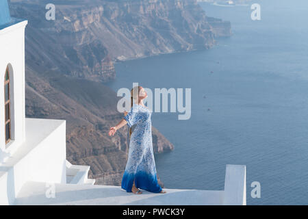 Donna felice in bianco e azzurro vestito godendo le vacanze sull'isola di Santorini, Grecia. Vista sulla Caldera e sul Mar Egeo da Imerovigli. Foto Stock