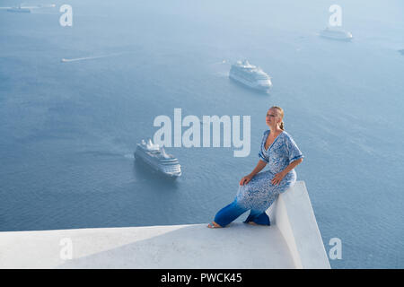 Donna felice in bianco e azzurro vestito godendo le vacanze sull'isola di Santorini, Grecia. Vista sulla Caldera e sul Mar Egeo da Imerovigli. Foto Stock