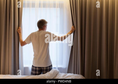 Persone di mezza età uomo caucasico apertura tende guardando attraverso la finestra in una camera di hotel Foto Stock