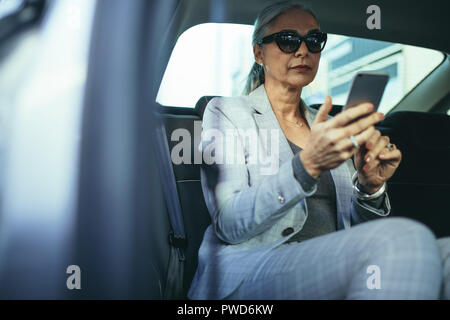 Elegante donna matura seduti sul sedile posteriore della sua automobile la lettura di un messaggio di testo sul telefono cellulare. Senior imprenditrice che viaggiano da un auto utilizzando smart phone. Foto Stock