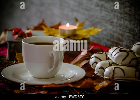 Tazza bianca con piattino con tè o caffè è sul tavolo, foglie di acero, accanto ad esso è una piastra con i cookie di bianco con strisce di cioccolato, un libro Foto Stock
