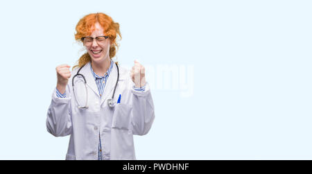 Giovani redhead donna che indossa uniformi medico entusiasta per il successo con le braccia sollevate celebrando la vittoria di sorridere. Vincitore del concetto. Foto Stock