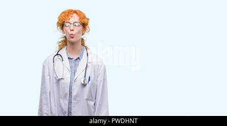 Giovani redhead donna che indossa medico rendendo uniforme il pesce faccia con labbra, pazzo e comico gesto. Espressione divertente. Foto Stock