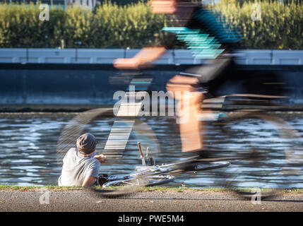 15 ottobre 2018, Assia, Frankfurt/Main: 15 ottobre 2018, Germania, Frankfurt am Main: Un ciclista si trova sulle rive del fiume Main e ha la sua bici proprio accanto ad esso. In primo piano un altro ciclista è a cavallo in direzione della bassa sun. Foto: Frank Rumpenhorst/dpa Foto Stock